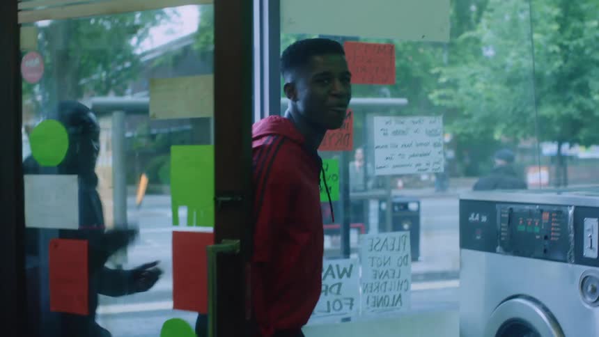 Three teenage boys open the door and smile and laugh as they walk into a laundry mat with signs on the glass