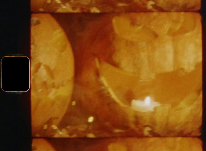 Panning handheld 8mm shot with horizontal camera movement of multiple jack o lanterns that are situated near each other surrounded by a light chain and brown oak leaves as candles burn inside of them while looking spooky and funny