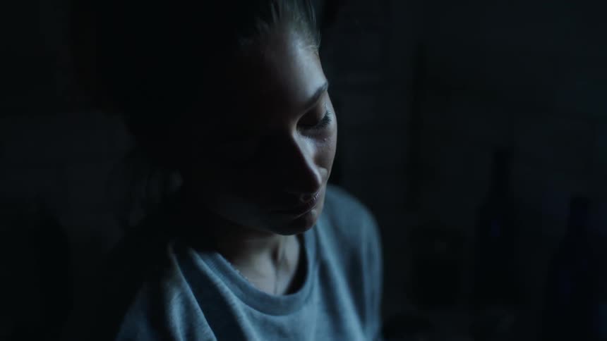Sad and lonely young woman in dark basement apartment or laundry room looking out the window and ruminating