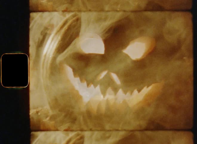 Handheld dynamic 8mm film footage close-up of the aggressive mouth of an angry Halloween pumpkin Jack O'Lantern glowing and smoking in the darkness as the camera zooms out revealing his evil eyes that cause a shocking surprise