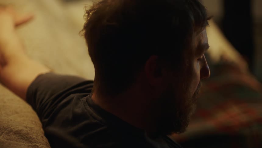 A bearded man sits on a couch while eating food with a fork