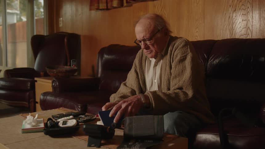 An older man wearing glasses sitting on the edge of his couch rubbing his hands together and then open a notebook to write in it