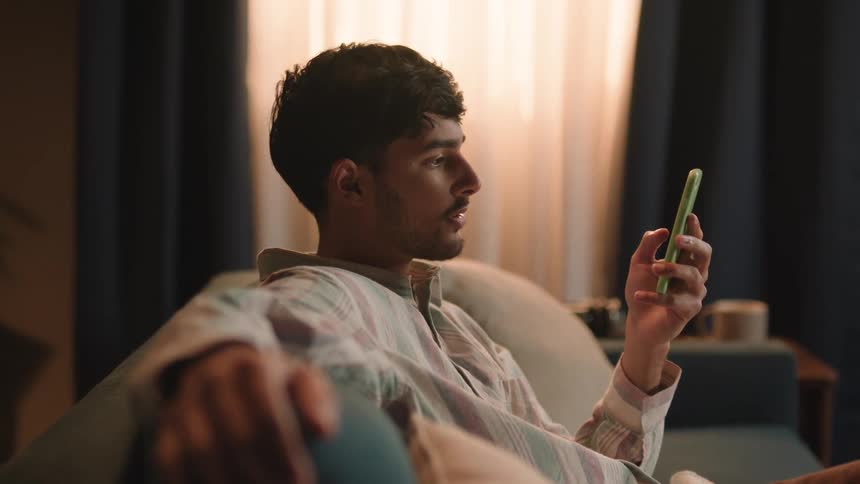 A man sitting on his couch scrolling on his cellphone with his left hand