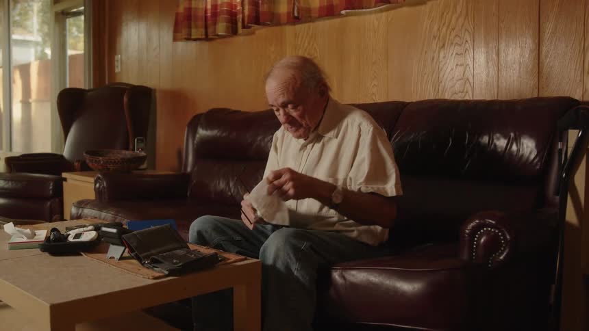 A man is sitting on his living room couch cleaning his eye glasses