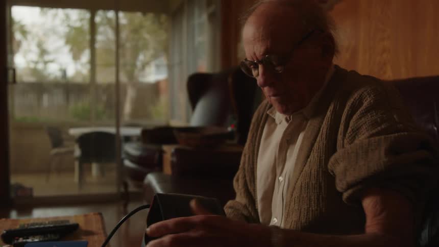 An older man raises his sweater sleeve and puts on a blood pressure cuff while sitting down