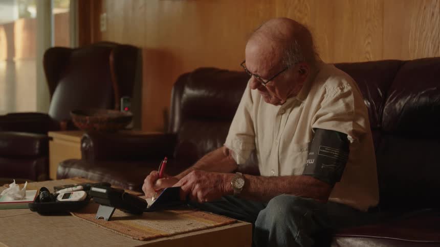 Elderly man sits on couch and writes while using blood pressure machine