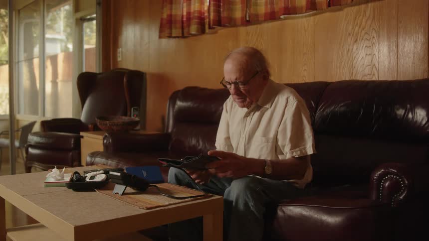 An older man wearing glasses sitting on his couch opening a blood pressure cuff