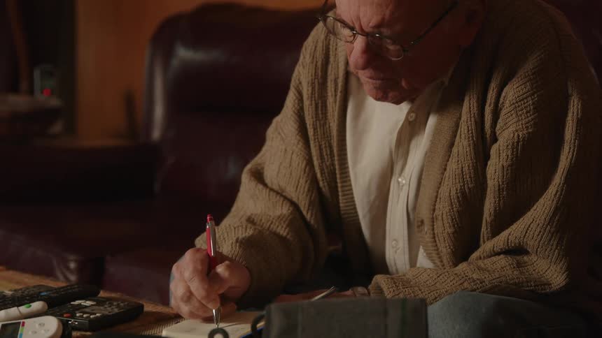 An older man sitting on his couch writing in a small notebook on the table in front of him