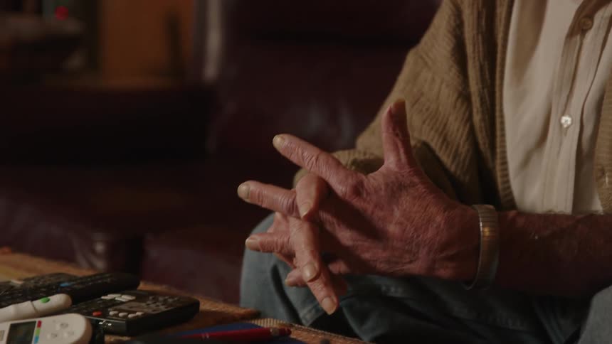 An old man sits on a couch while the camera pans to show his hands and an electronic device