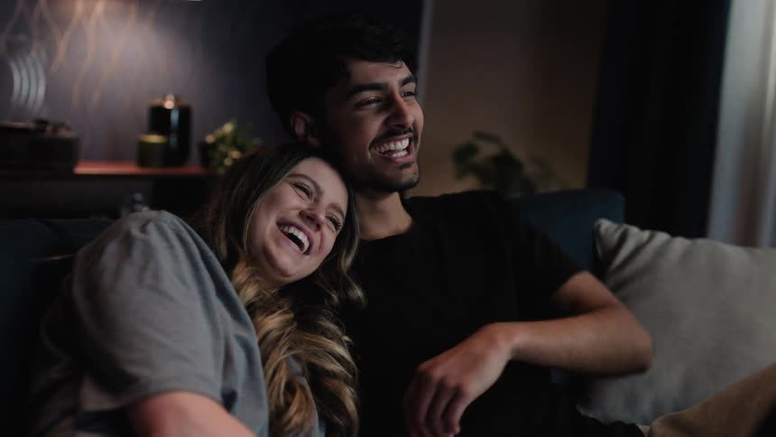 A man and woman sit on a couch leaning against each other laughing and smiling and pointing ahead of them
