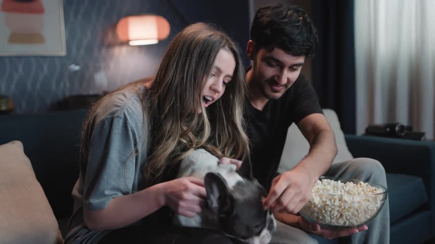 A man eats popcorn while a woman pets the dog