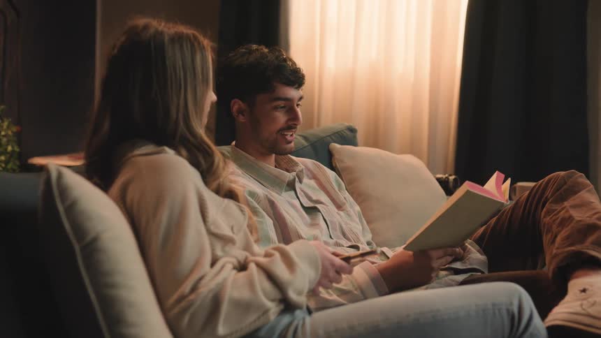 Young happy couple sitting on a couch reading a book togheter at home