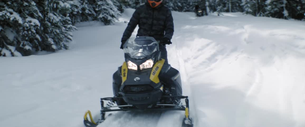A person on a snowmobile driving through a snowy forest as another person drives a snowmobile behind them