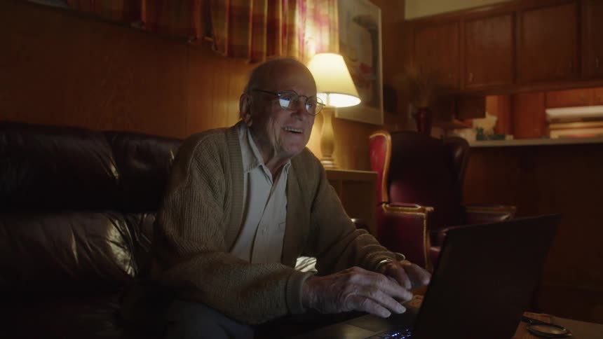 An older elderly man is sitting down and is on his laptop