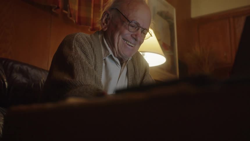 Elderly man typing on a laptop in the living room smiles and laughs