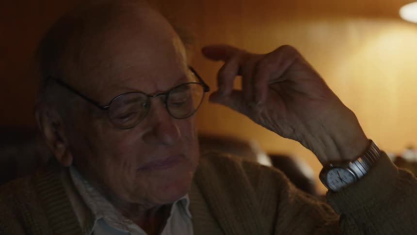 A man is sitting in his living room holding his glasses in his hand and thinking for a moment before he puts them on his face
