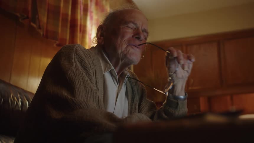 An elderly man contemplates takes off his glasses then smiles and laughs to himself