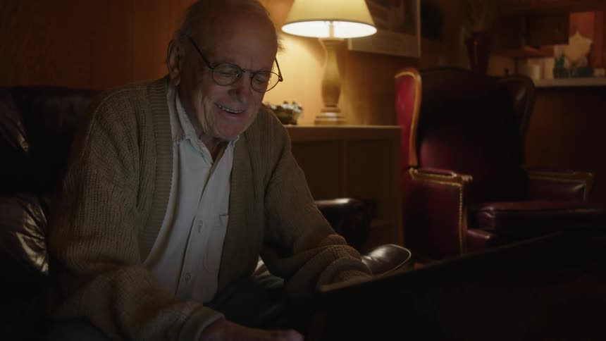 An older elderly man is sitting on the couch and looking at his computer