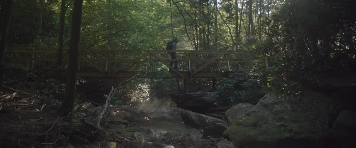 Man wearing a backpack walks over a small bridge in the woods