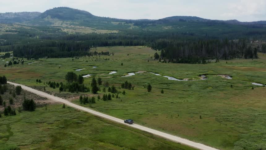 A truck travels on a rural road as trees are scattered in the field with forests and mountains in the distance
