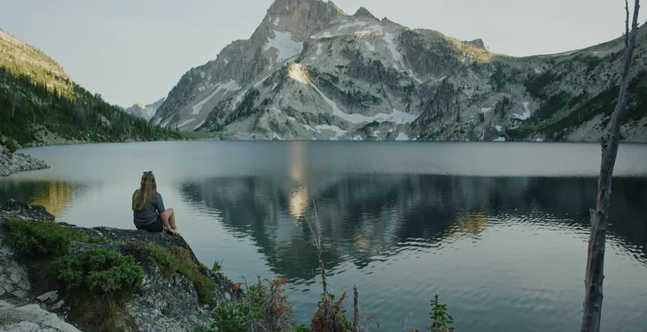 Woman sits on shore looking across lake reflecting nearby mountains
