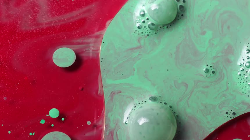 A red substance moves as a green splashes over the surface and bubbles float as the colors blend and green dots float over the red area