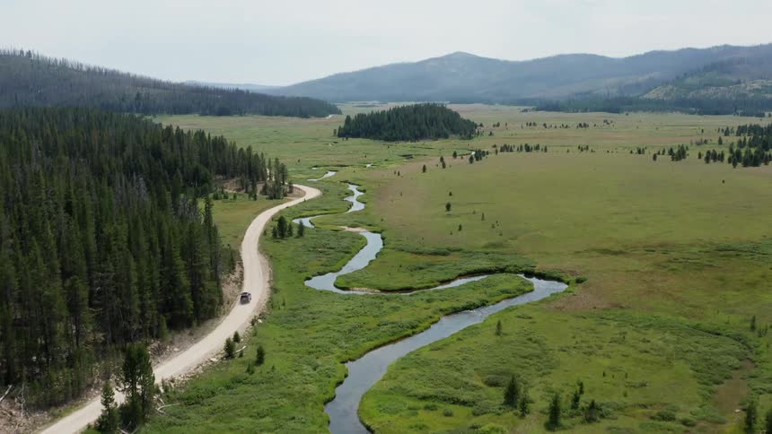 An aerial view of a grassy land with a dirt road and a car traveling on it and mountains in the distance