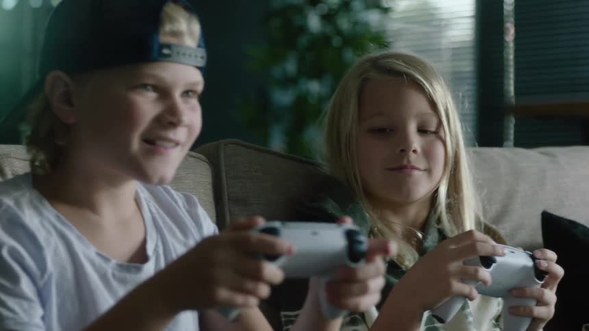 Brother and sister play console video games with controlers on the couch at home