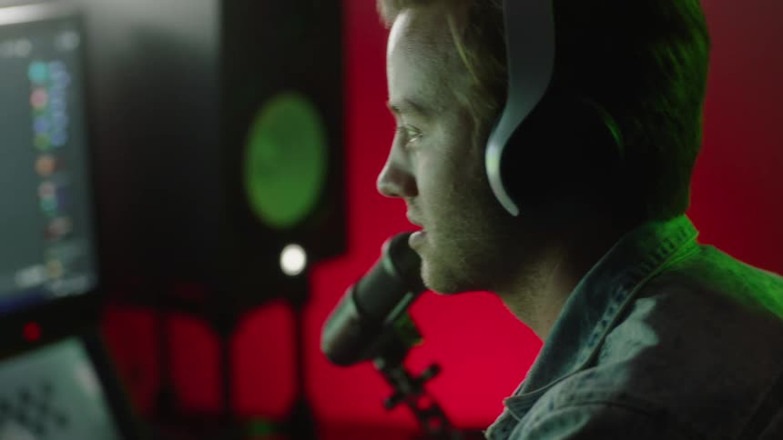 A close up of a mans face with headphones on his head while he talks in to a microphone in front of his computer