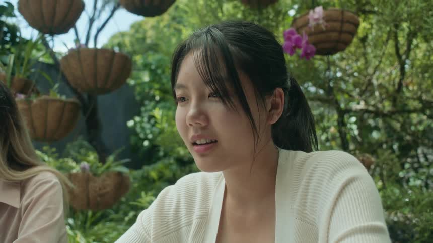 A woman sitting outside looking contemplatively next to another woman sitting under hanging plants