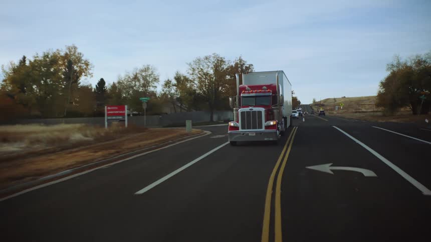 A red semi truck is driving down the street and surrounded by trees in the fall