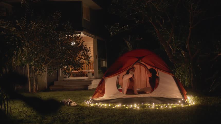 Two young women are camping in a tent in the backyard of a twostory house