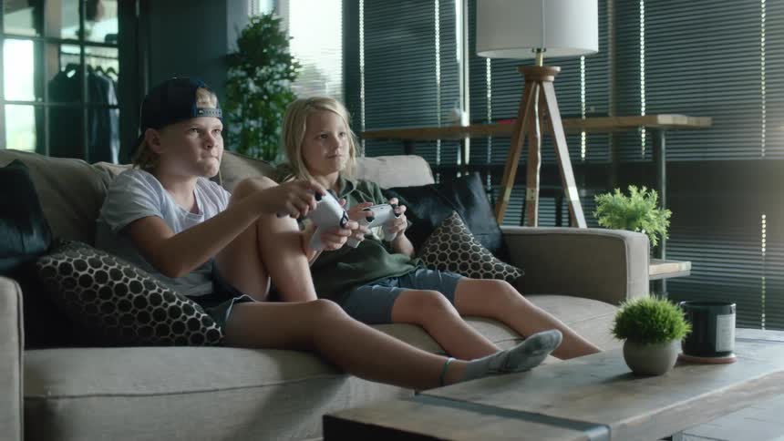 A pair of children play video games together on a sofa during the day