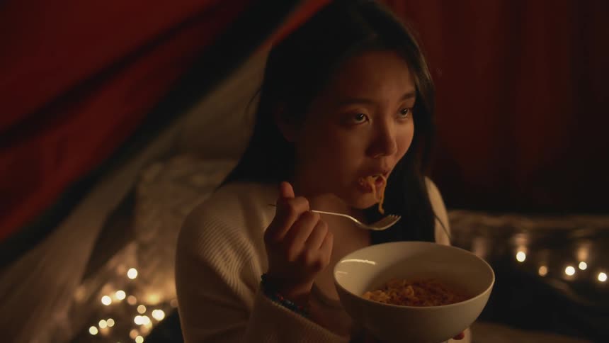 Young woman sitting in a tent with friends eating a bowl of noodles at nighttime