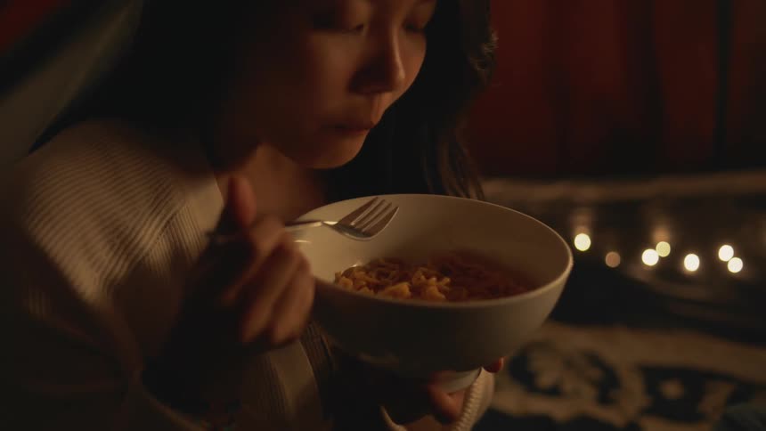 A close up of a bowl of noodles in a womans hand while she eats with a fork