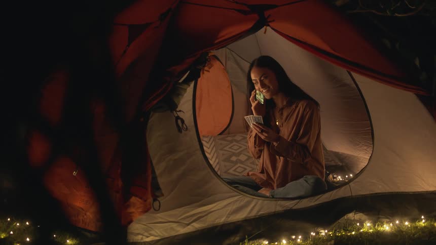 Woman with dark hair shirt and jeans sits in tent talking on smartphone