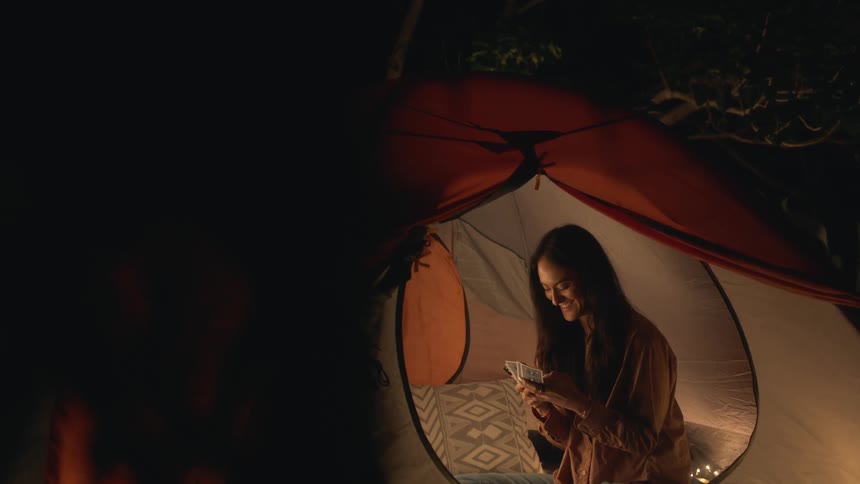 A woman is playing a game of cards inside a camping tent with someone during the evening