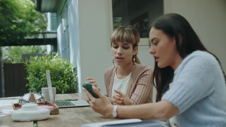 Two women sitting at a table outside one holds a cellphone and leans over to the other pointing at her phone