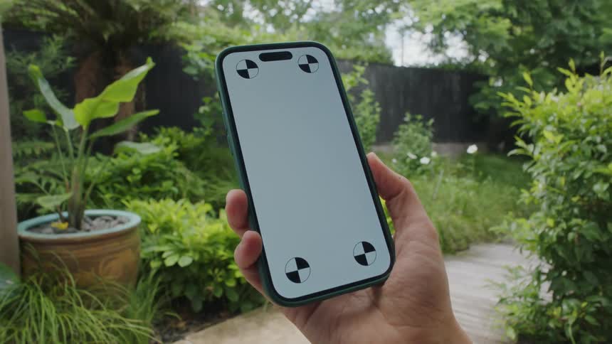 A man in a back yard lifts up a cell phone with a white screen with circles in each corner