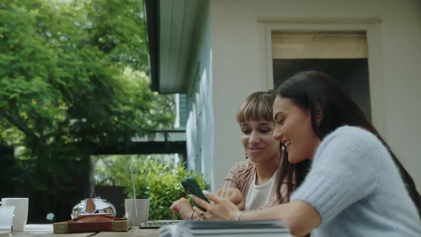 Two women sit at a table outside one picks up her phone and shows it to the other as they both smile and laugh