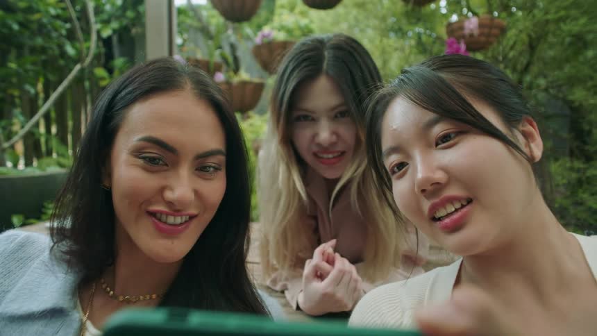 A group of women sitting outdoors laugh while looking at a smartphone together
