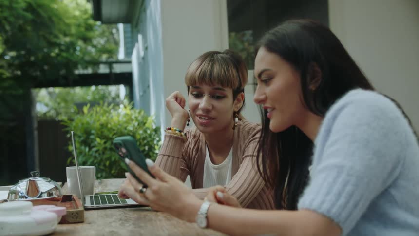 Two women sitting at a table outside leaning over to one another looking at the phone in the one womans hands