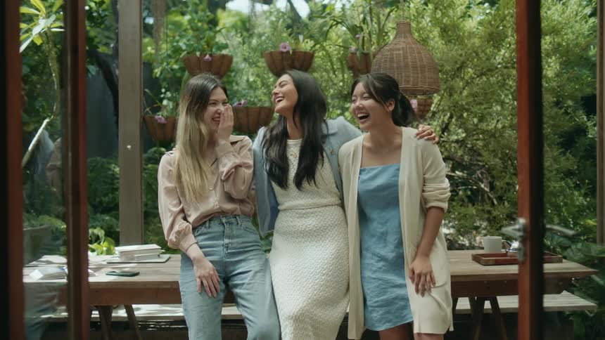 Three teenage girls are standing outside leaning on a table and laughing as the girl in the middle has her arms around the other two girls