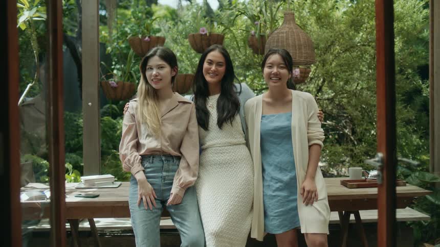 Three teenage girls are standing outside leaning on a table and laughing as the girl in the middle has her arms around the other two girls