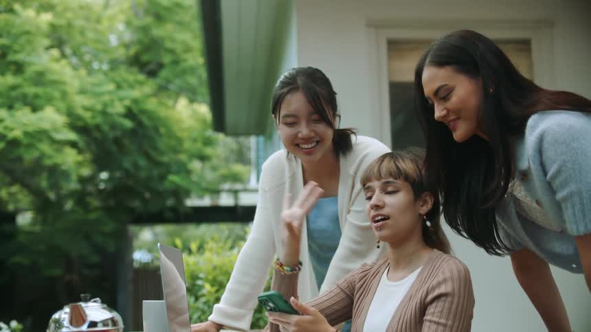 Three woman standing around each other outside while they look at a phone in the persons hands who is sitting