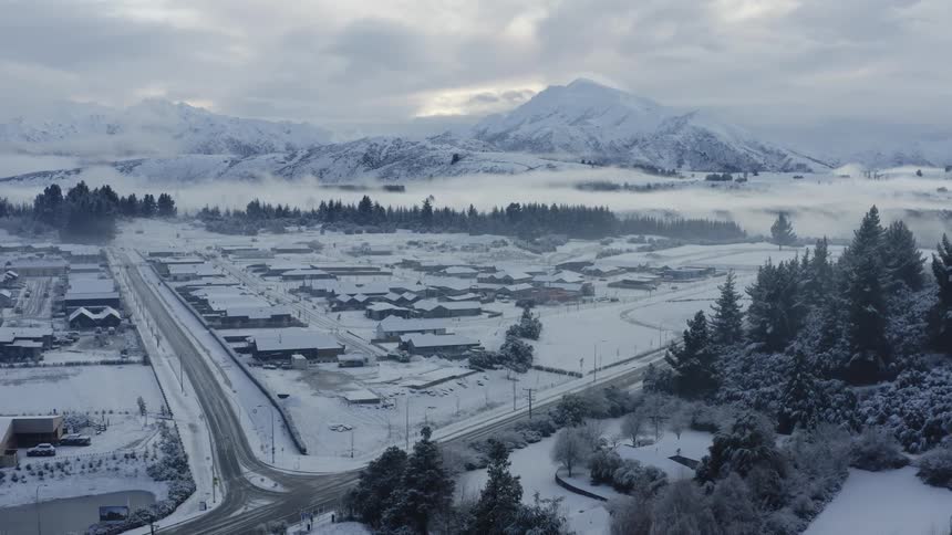 Snow covered town sits between mountains and forest under an overcast sky