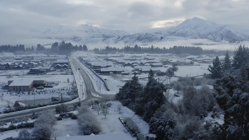 Vehicle drives along road away from intersection with road passing through snow covered town bordered by woods and mountains under an overcast sky