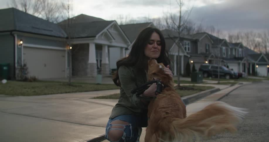 A woman kneeling down petting a dog smiling and laughing near a neighborhood sidewalk