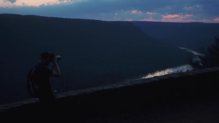 Photographer standing near wall takes picture of river curving around mountains under a red sky