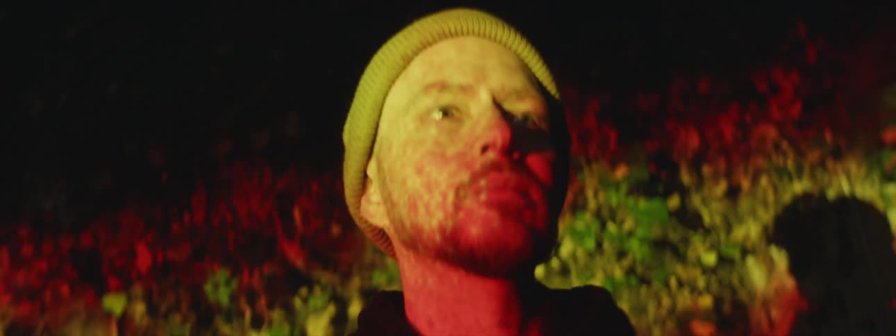 A man is wearing a knit hat as colored lights move across his face and on the wall behind him